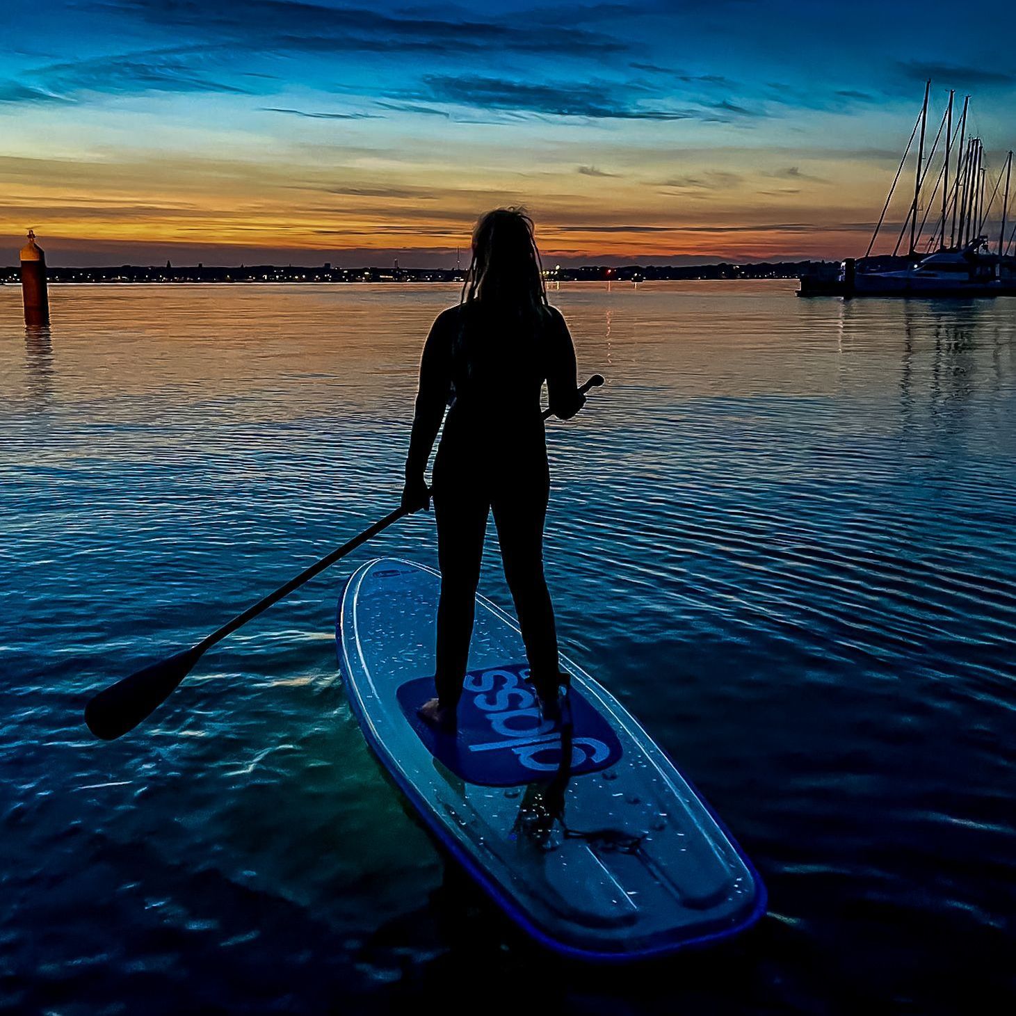 Eine Frau steht auf einem glassUP im Wasser vor traumhafter Hafenkulisse im Sonnenuntergang.