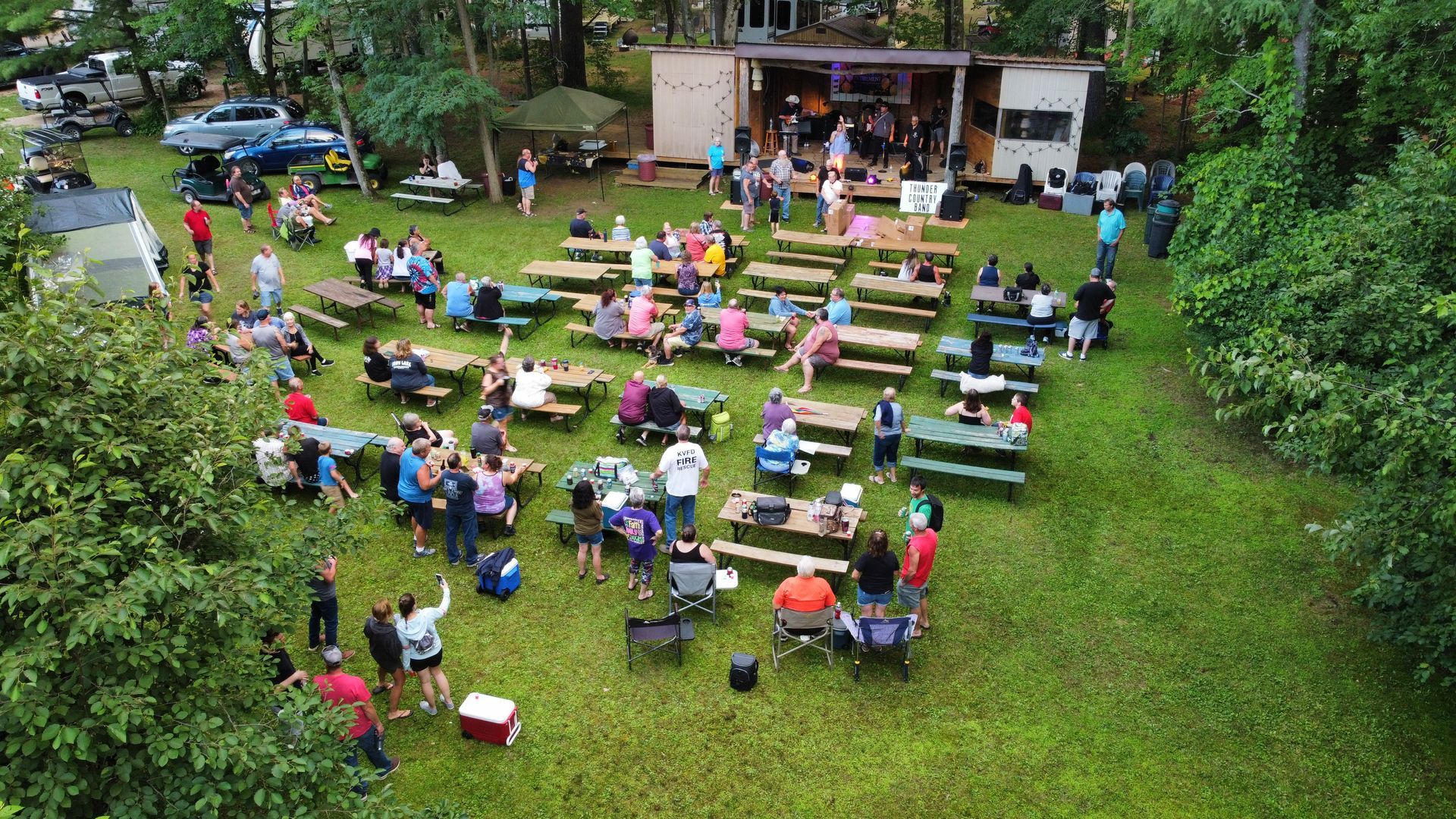 An aerial view of a large group of people sitting at picnic tables in a grassy field.