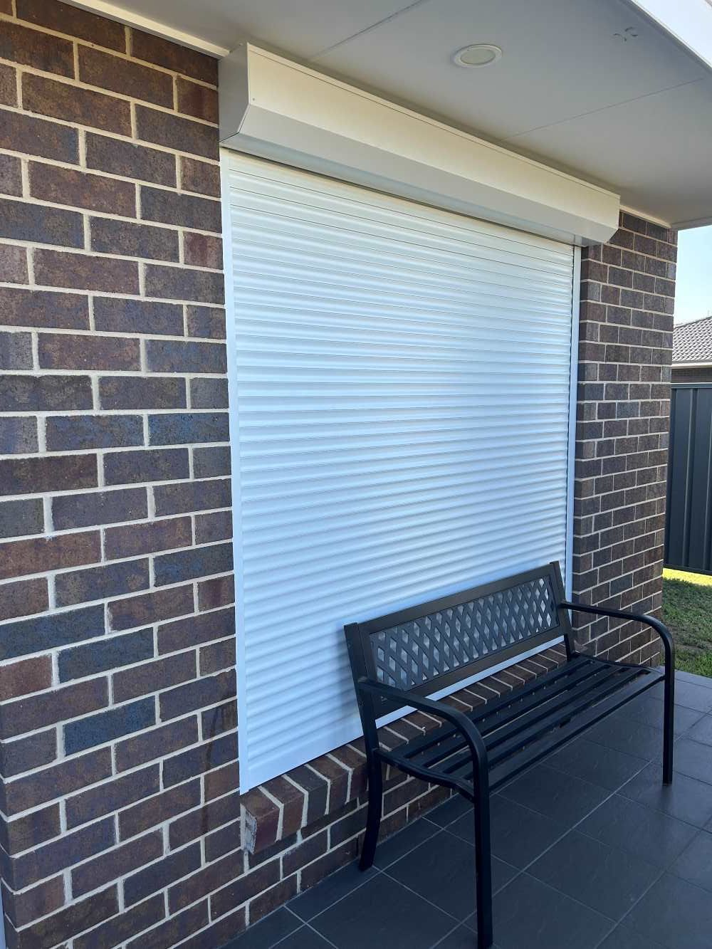 Black Bench Is Positioned In Front Of A Window With A White Roller Shutter — Maxview Blinds and Shutters in Beresfield, NSW