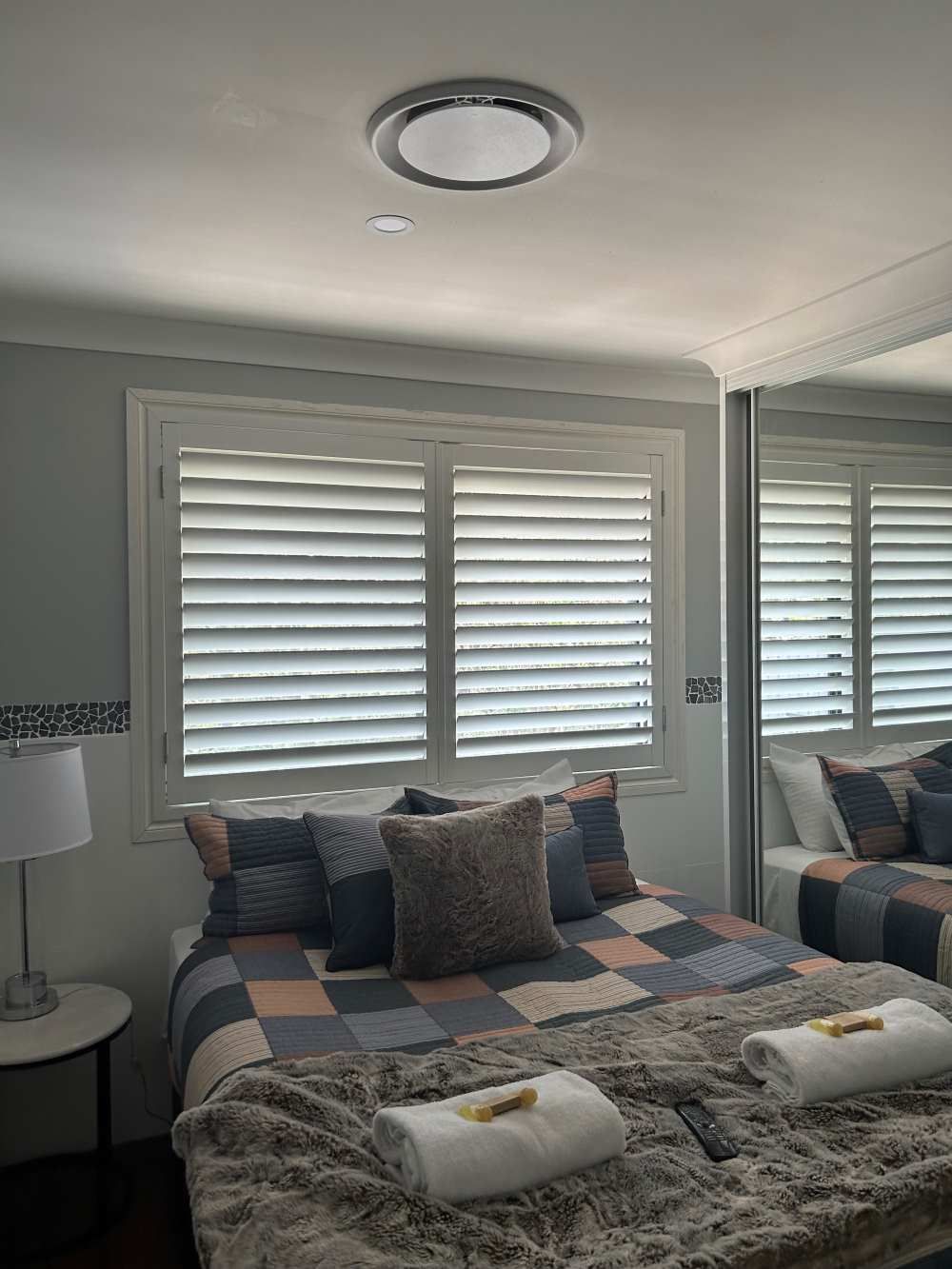 Bedroom Furnished With A Bed, A Lamp, A Mirror, And Window Shutters — Maxview Blinds and Shutters in Beresfield, NSW