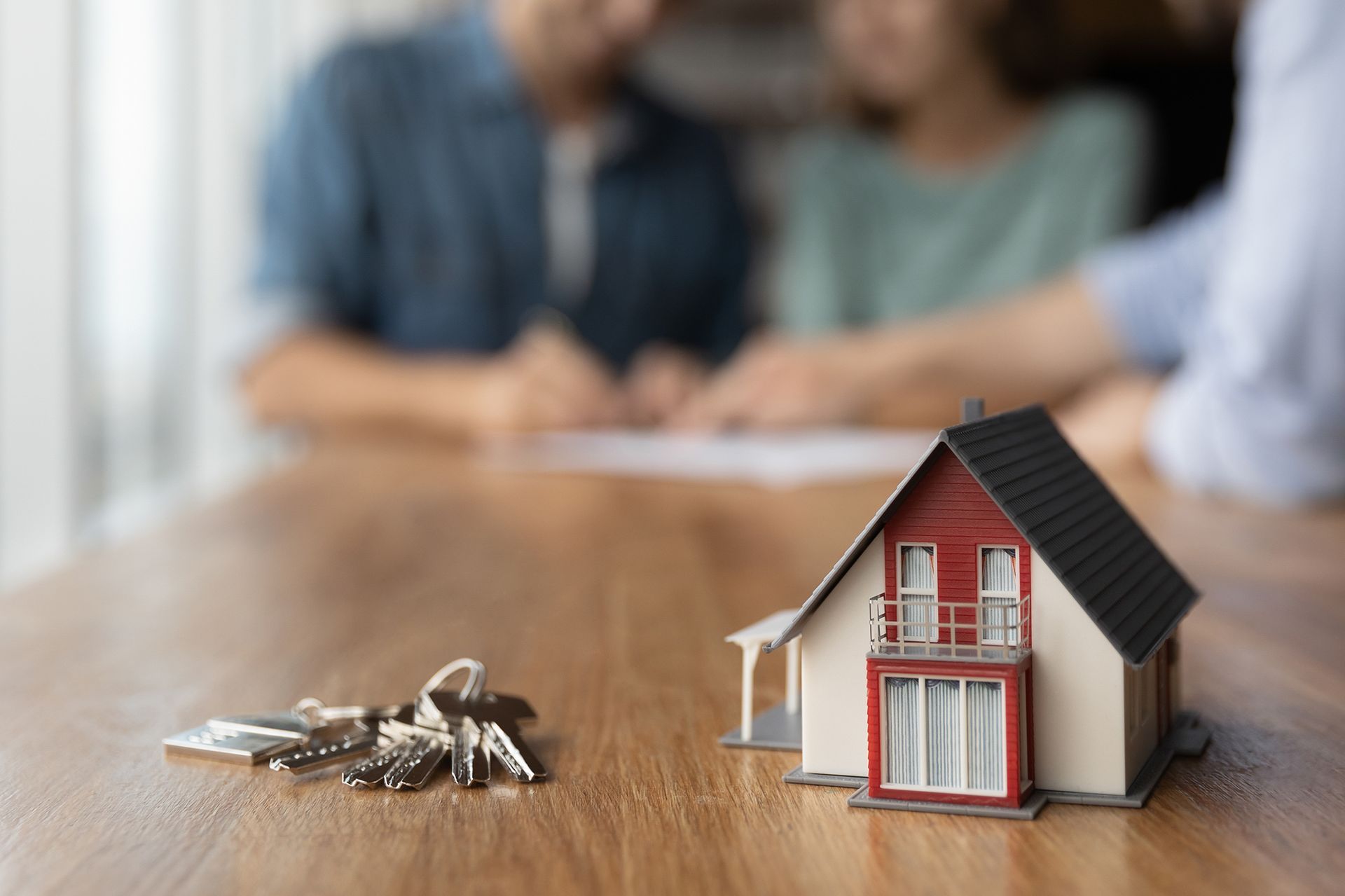 Miniature house with keys on a table as a couple signs paperwork with a blurred advisor.
