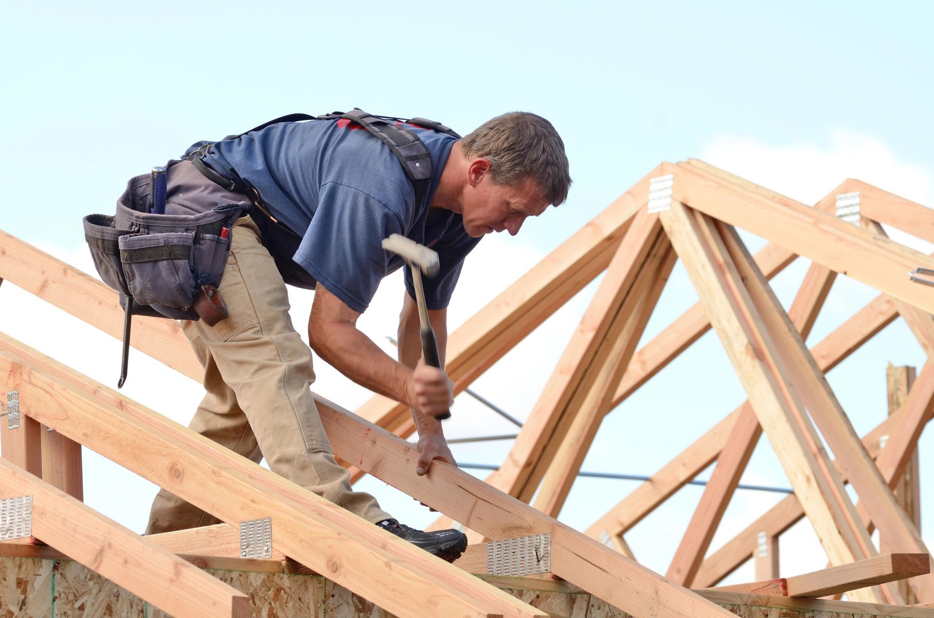 An Australian carpenter with a builders license on a timber roof frame, securing beams under a clear blue sky.