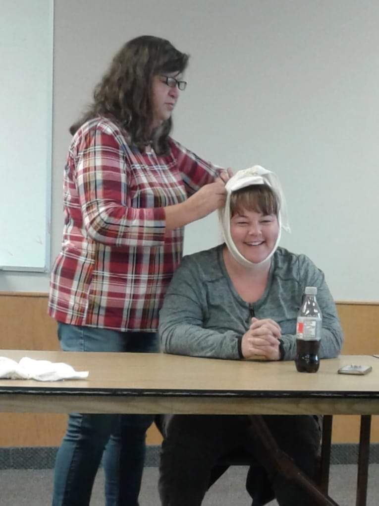 A lady pinning something on a lady siting on a chair at a table.  Big smile.