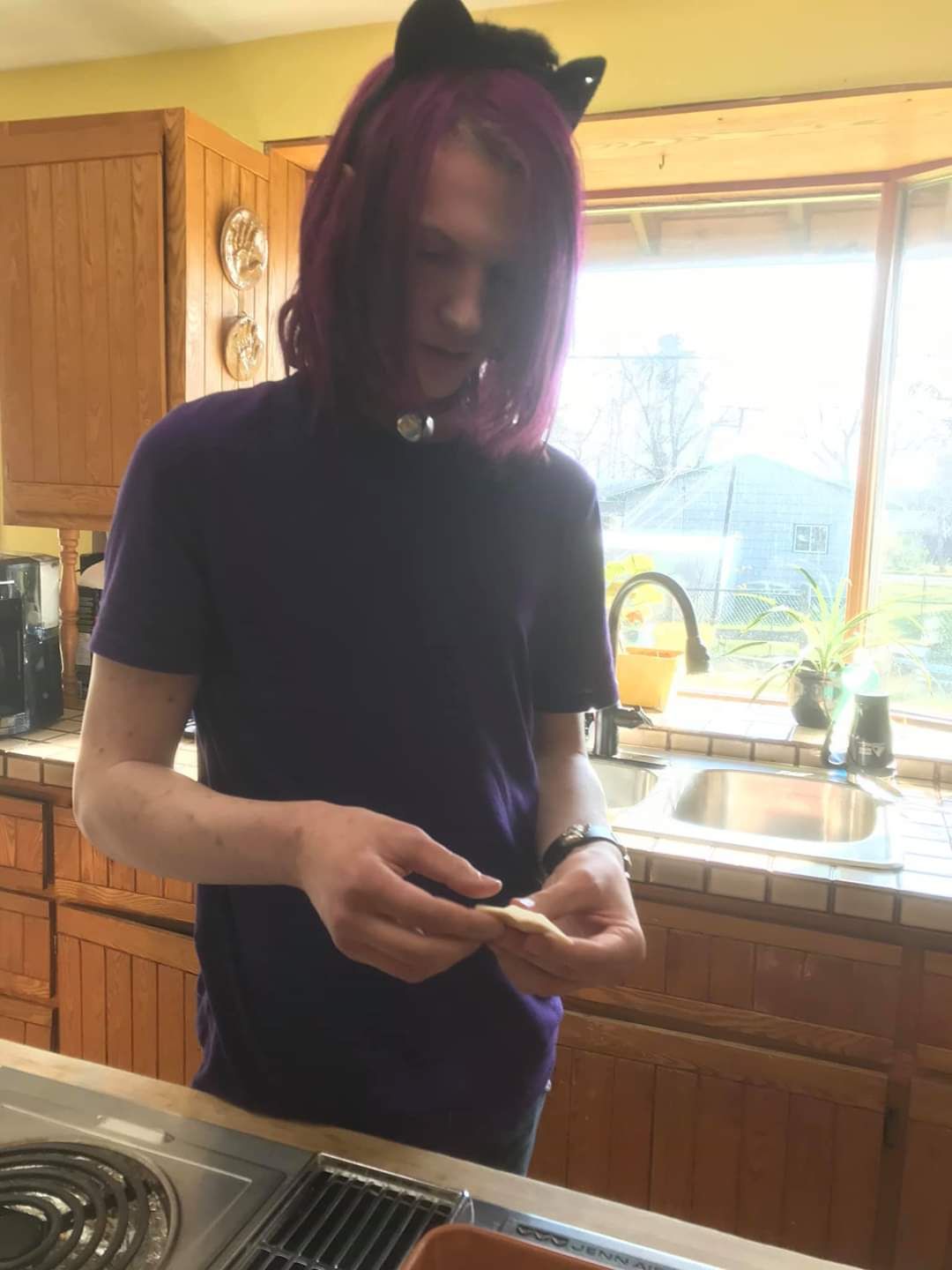 A man with purple hair and cat ears is standing in a kitchen looking at his phone.