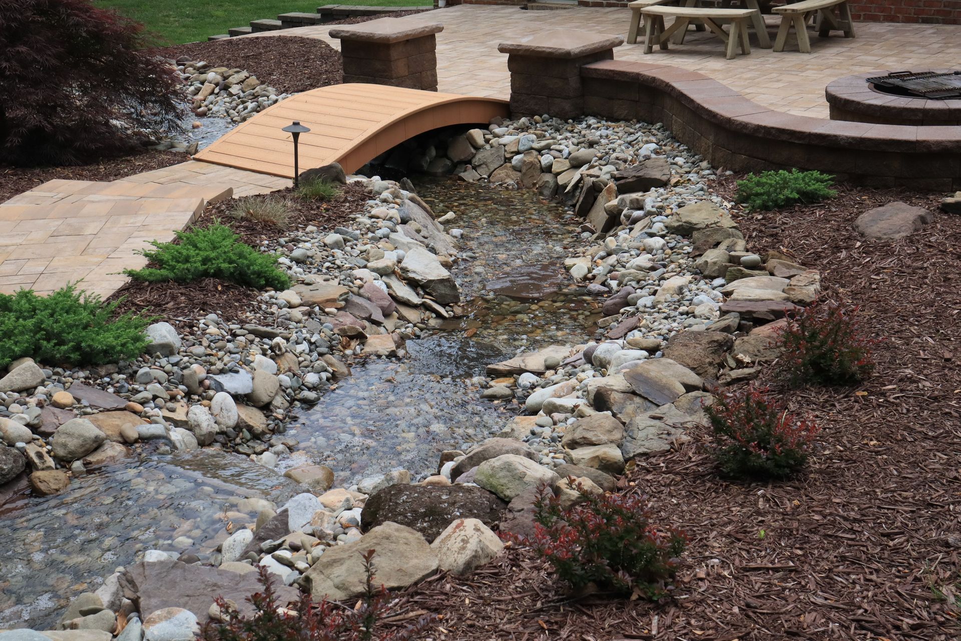 A decorative backyard stream with a small wooden bridge, surrounded by rocks and landscaping.