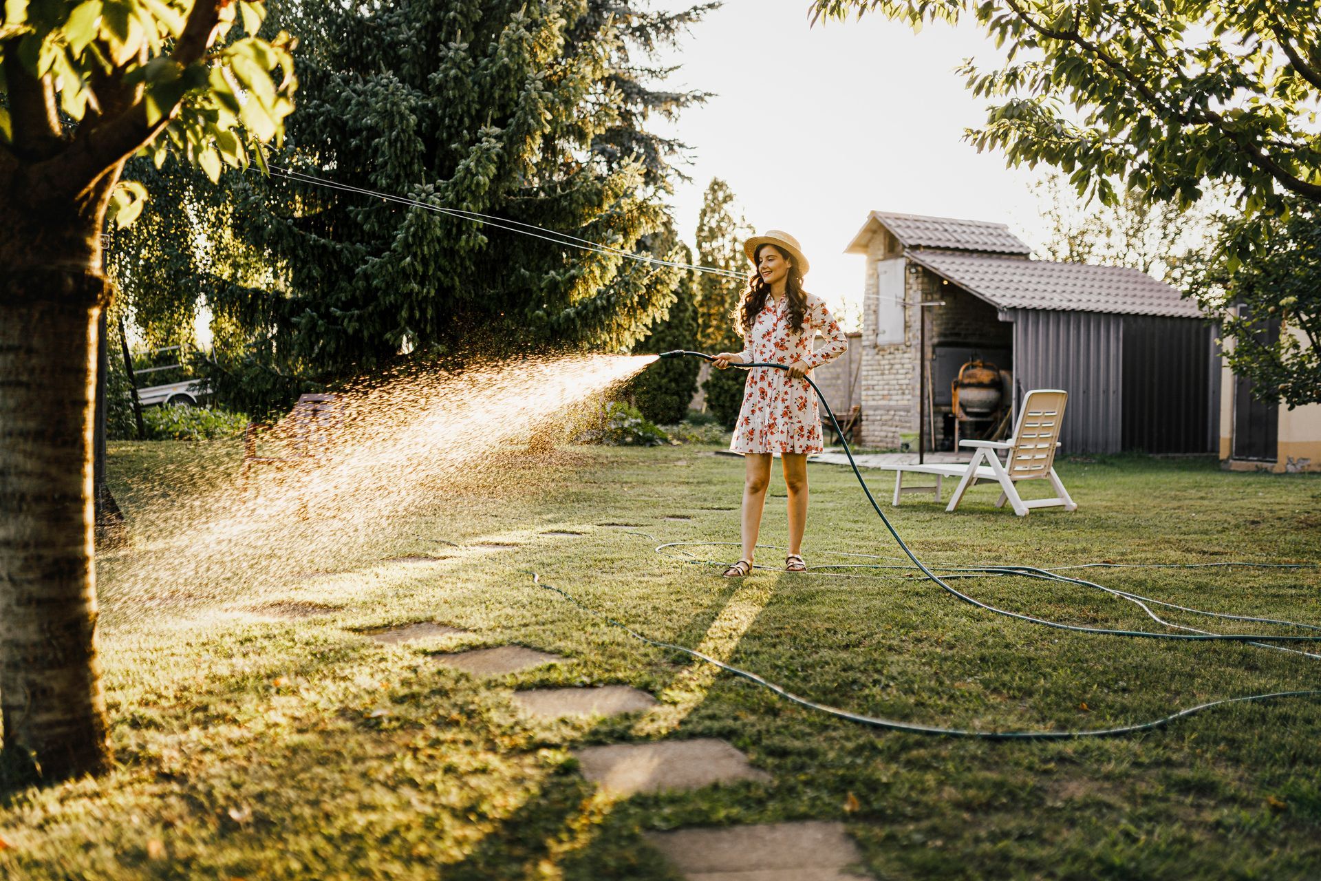 Woman in floral dress watering a grassy backyard with a hose in the sunlight.