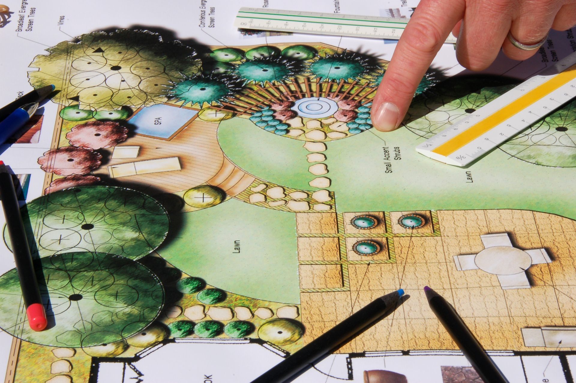 Hand pointing to a colorful landscape design plan with trees, a fountain, and pathways.
