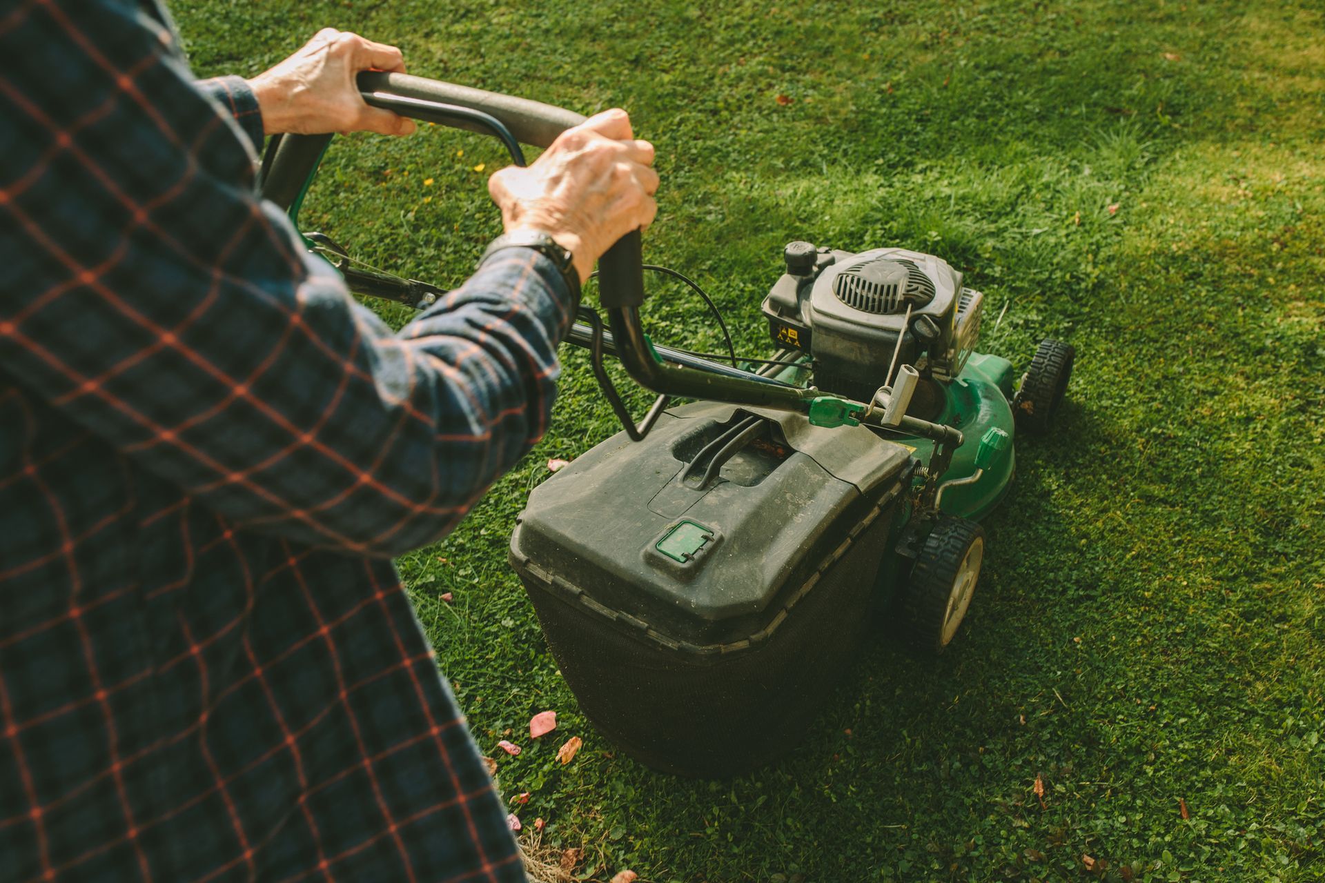Man in plaid shirt mowing green lawn with a gas-powered mower.