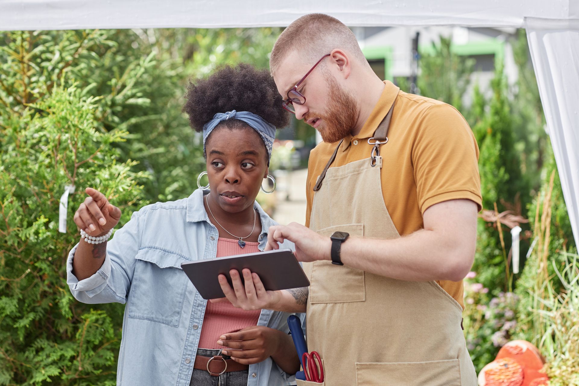 Woman pointing, looking at a tablet with a man in an apron. They are outside, likely at a nursery, discussing plants.