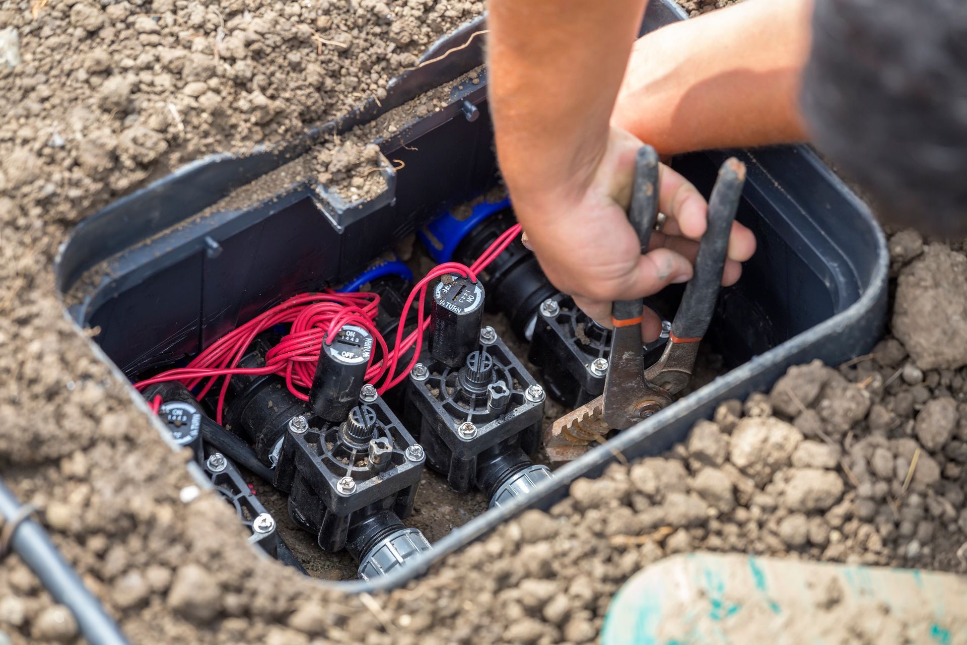 Person using pliers to work on irrigation system valves in an in-ground valve box. Red wires visible.