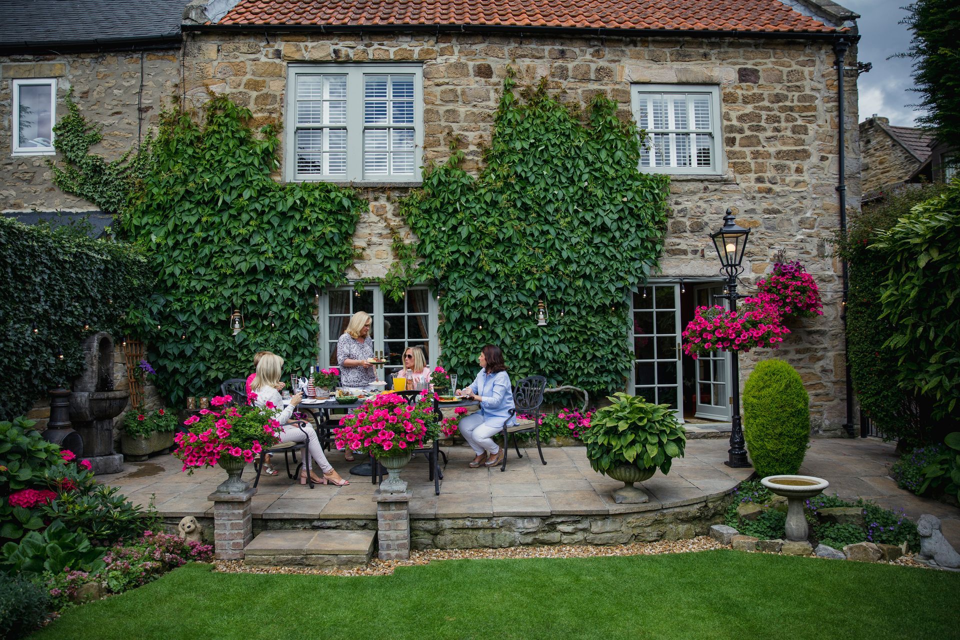 Four women at a stone patio table, enjoying drinks at a brick house with ivy and flower pots.