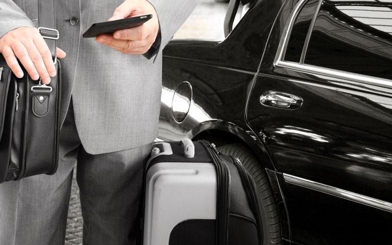 Person in suit holding phone and briefcase, standing next to a black car and suitcase.