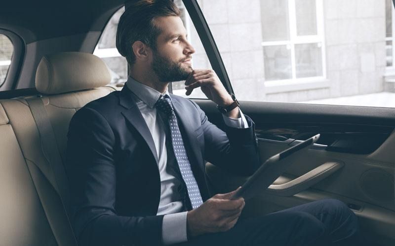 Man in a suit in the backseat of a car, looking out the window, holding a tablet.