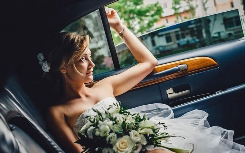 Bride in white strapless dress, holding bouquet, looking out car window, arm raised.