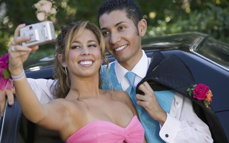 Couple taking selfie; woman in pink dress, man in suit with a car in the background.