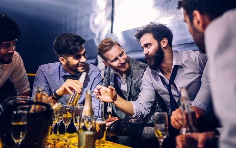 Men at a table, some holding drinks, in a dimly lit setting, smiling and talking.