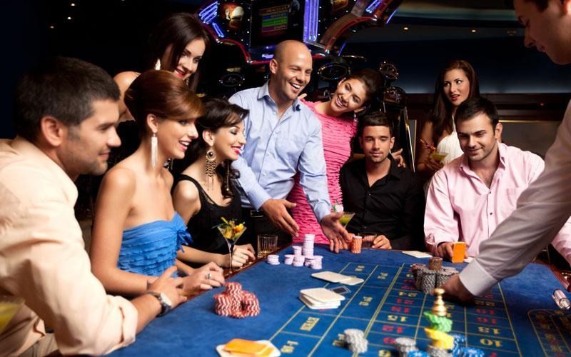 People gathered around a casino roulette table; some smiling, placing bets. Blue felt, bright lights.