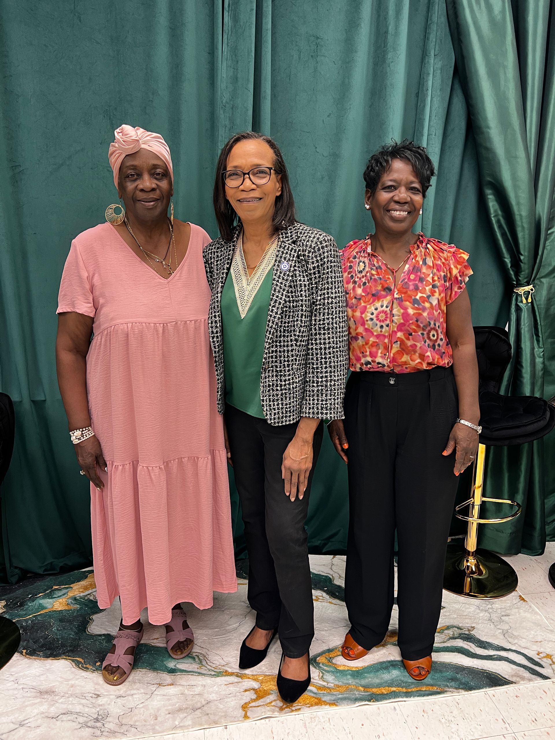 Pulaski County NAACP President Cynthia Andry wears a pink dress and head wrap, Yvonne Reeves-Chong wears a black and white jacket, and  Lois Hollins wears a floral shirt while posing in front of a green curtain.