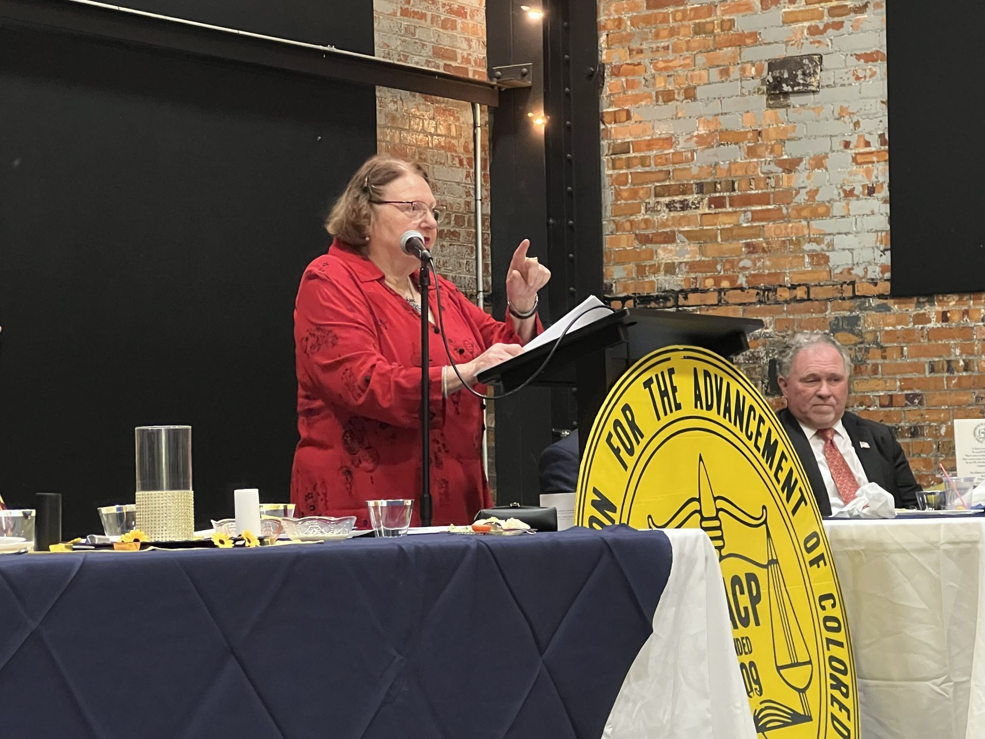 Woman in red top speaking at a lectern, man in suit seated behind a table with a sign that reads 