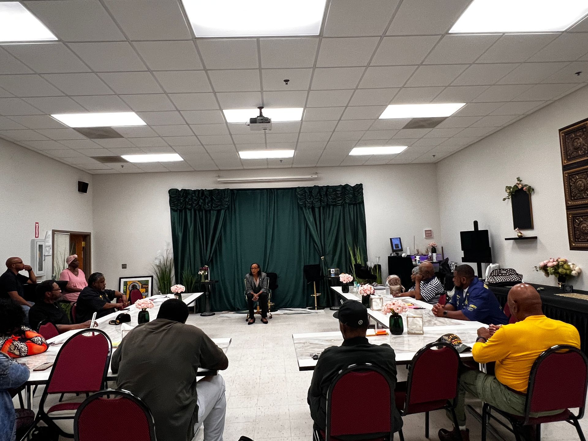 People seated at tables in a brightly lit room, looking toward a person speaking in front of a curtain.