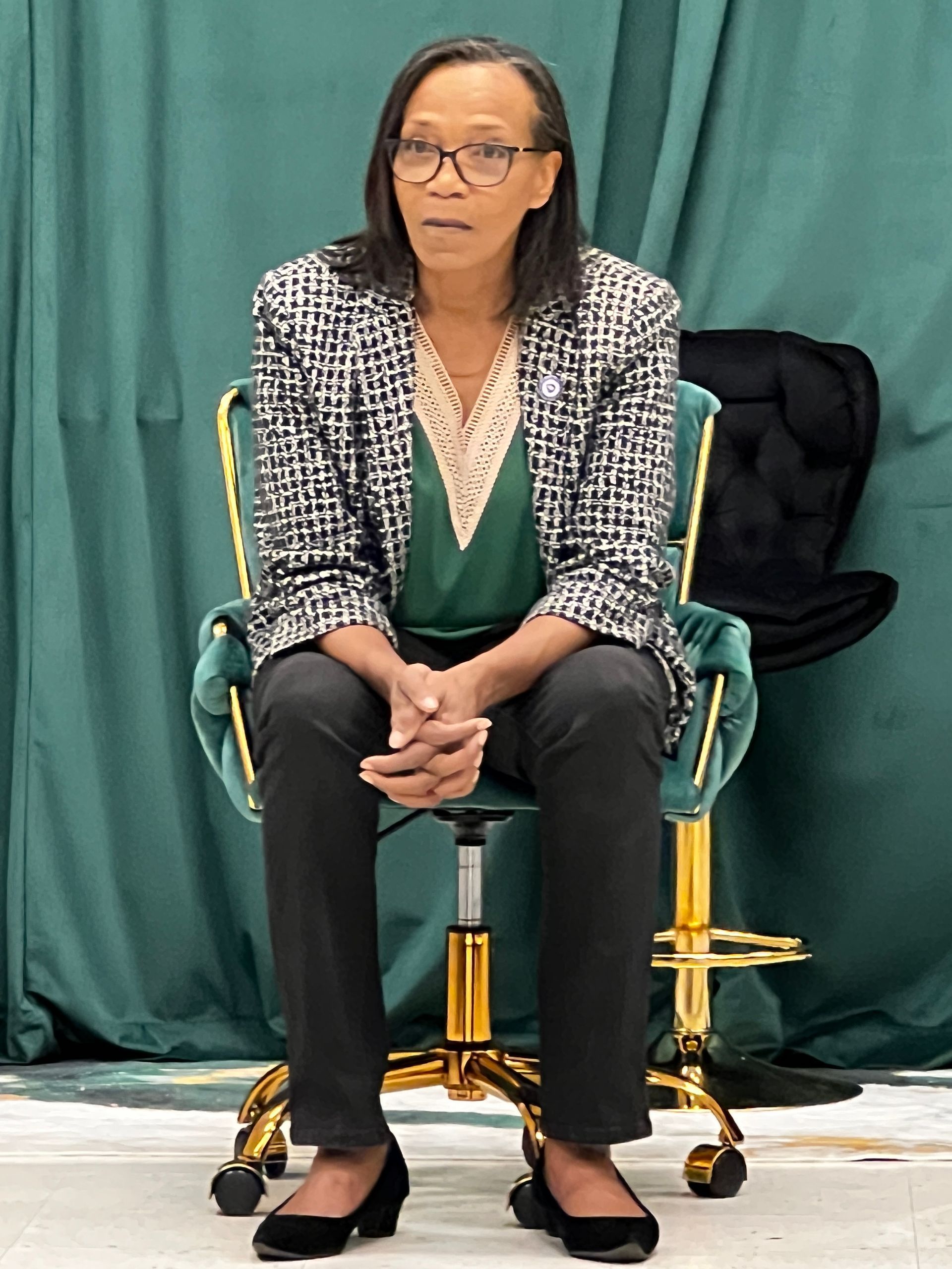 Yvonne Reeves-Chong, a black woman in a green shirt and white and black checked jacket, sits leaning forward while speaking to the Pulaski County NAACP Branch.