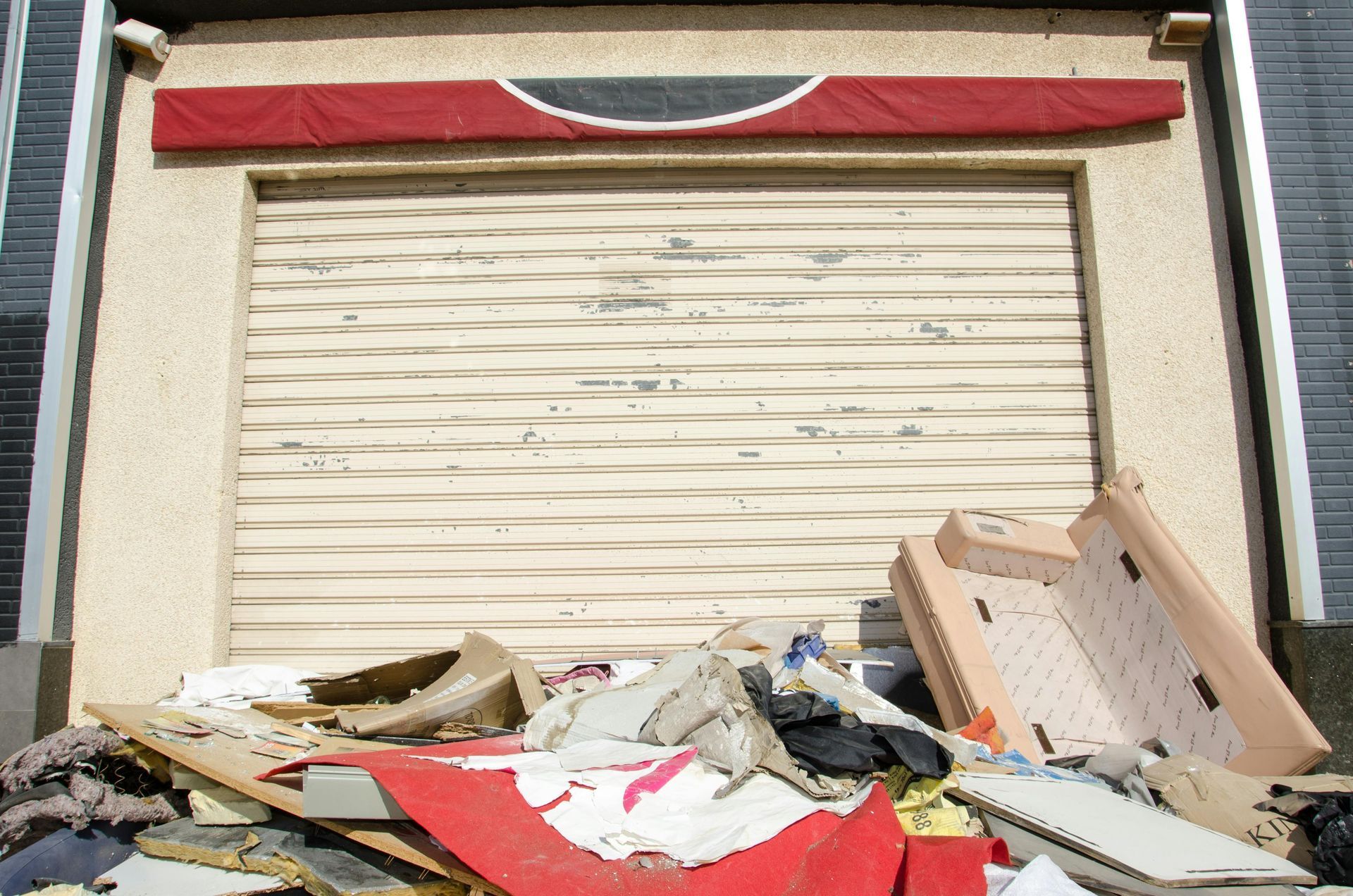 Closed shopfront with beige roll-down gate and debris piled in front. Red awning detail above.