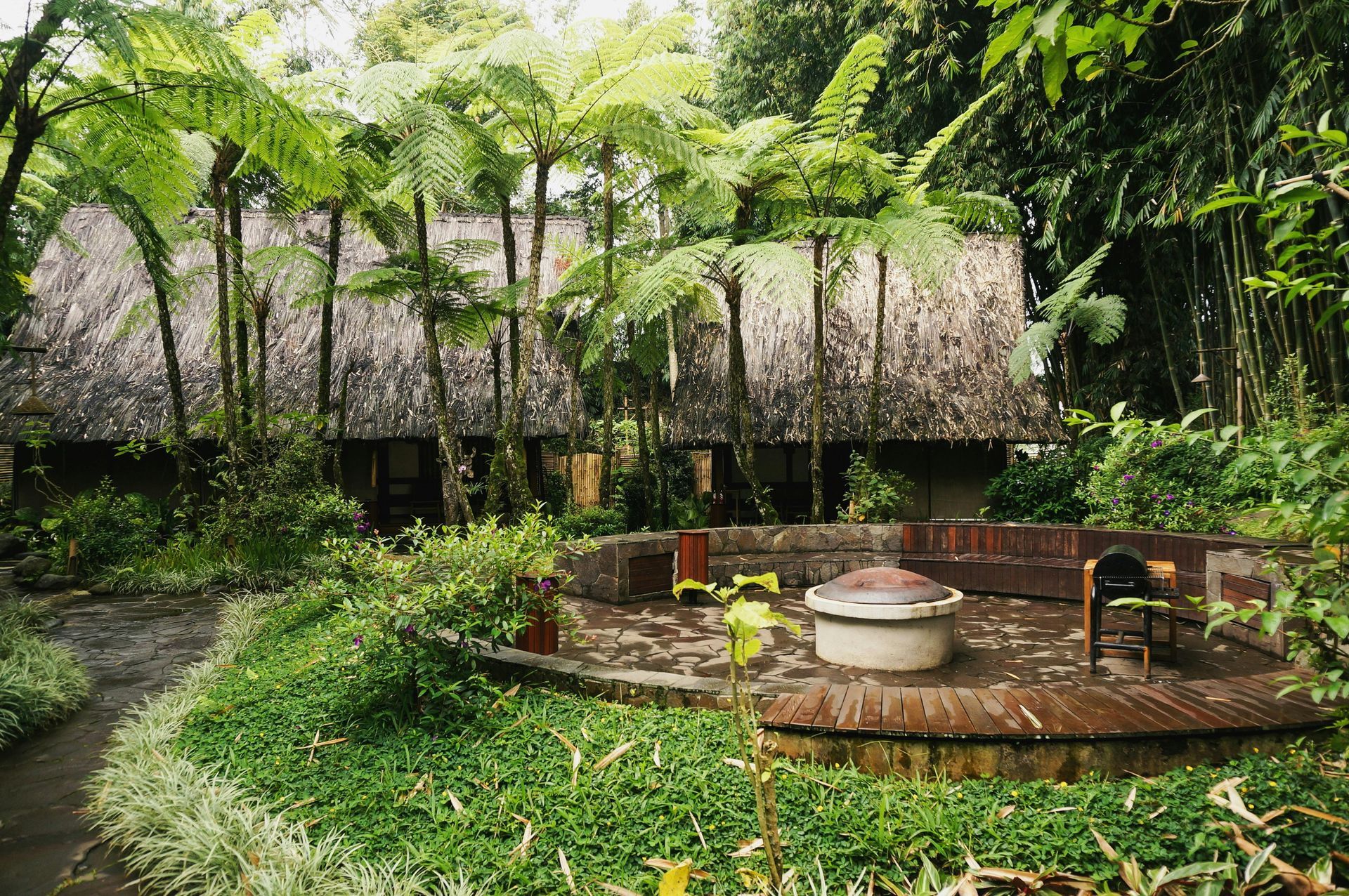 A tropical lodge with a thatched roof, surrounded by lush green plants and a circular brick seating area.