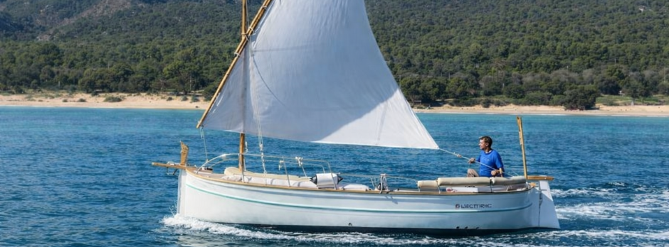 Llaut eléctrico navegando por la costa de Mallorca con vela latina y paisaje mediterráneo de fondo