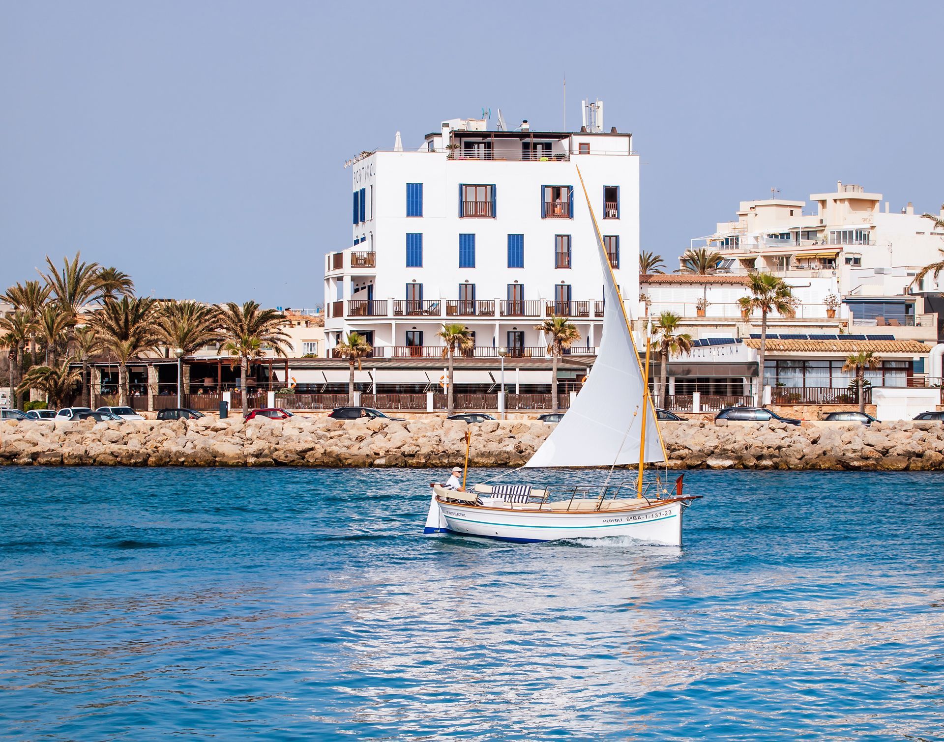 Un llaut navega por el mar, con un edificio moderno al fondo, creando un contraste entre lo tradicional y lo urbano.