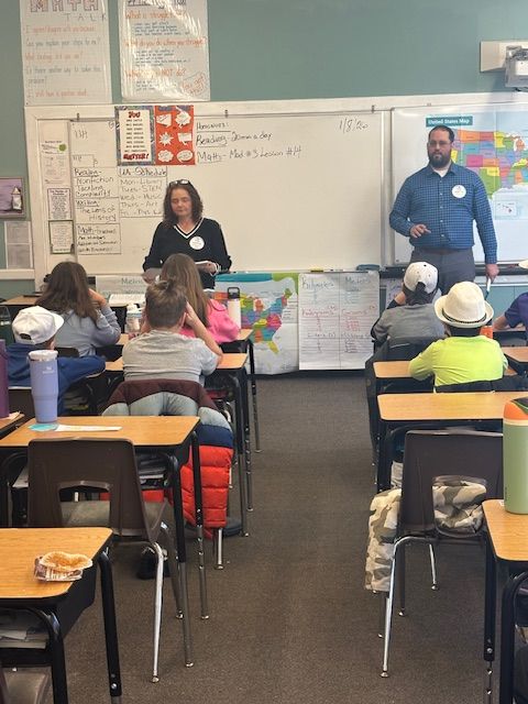 Two adults stand in a classroom, presenting to a group of students seated at desks facing a whiteboard and maps.