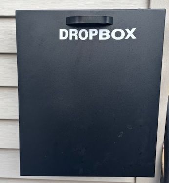 A black, wall-mounted metal dropbox with a handle, mounted on light-colored horizontal siding.