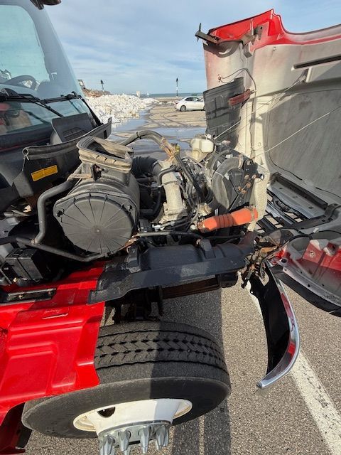 A red semi-truck with its hood tilted forward, exposing the engine bay while parked near a shoreline.