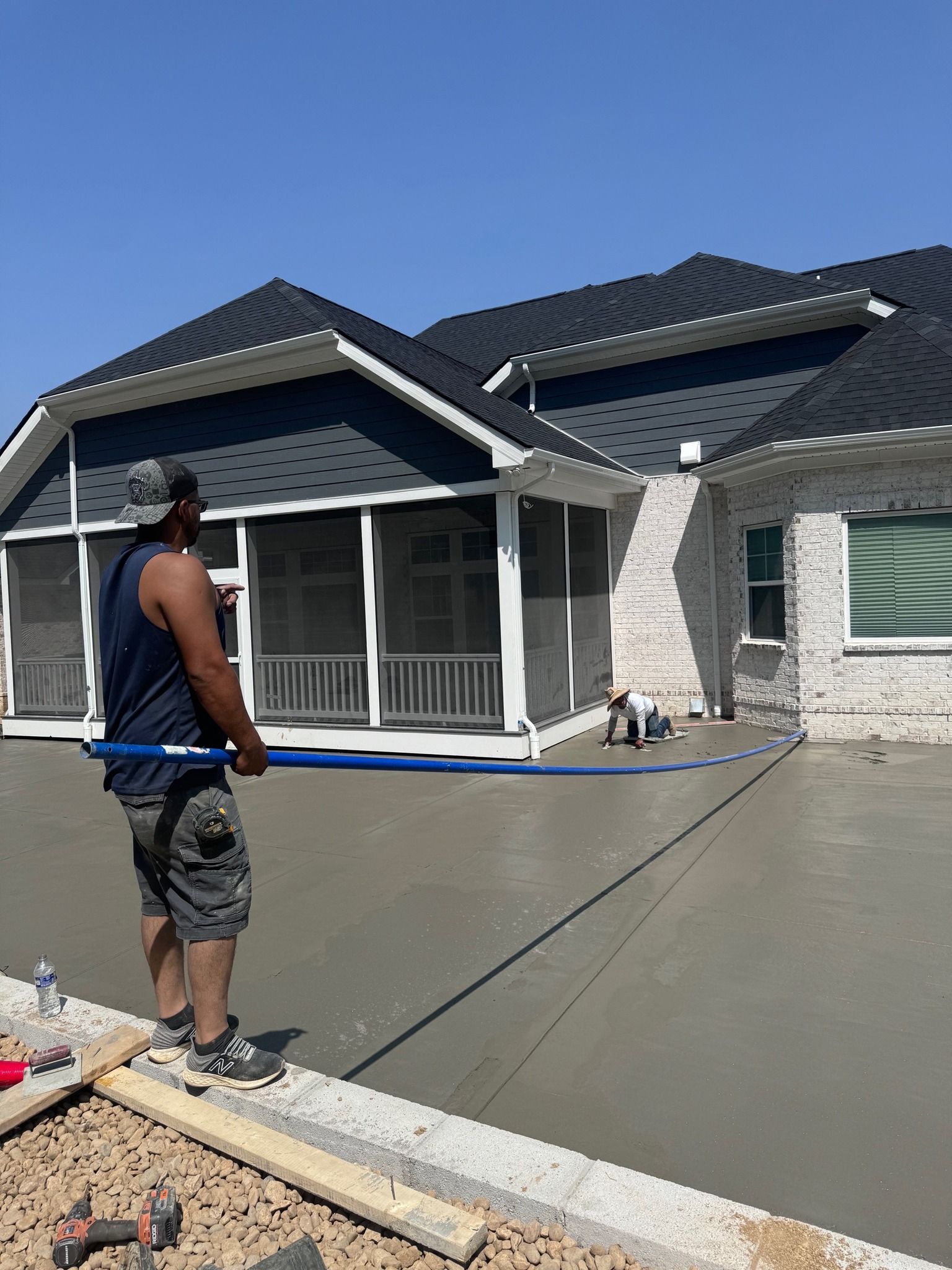 Man leveling wet concrete patio with a long tool; another person works nearby. Building in background.