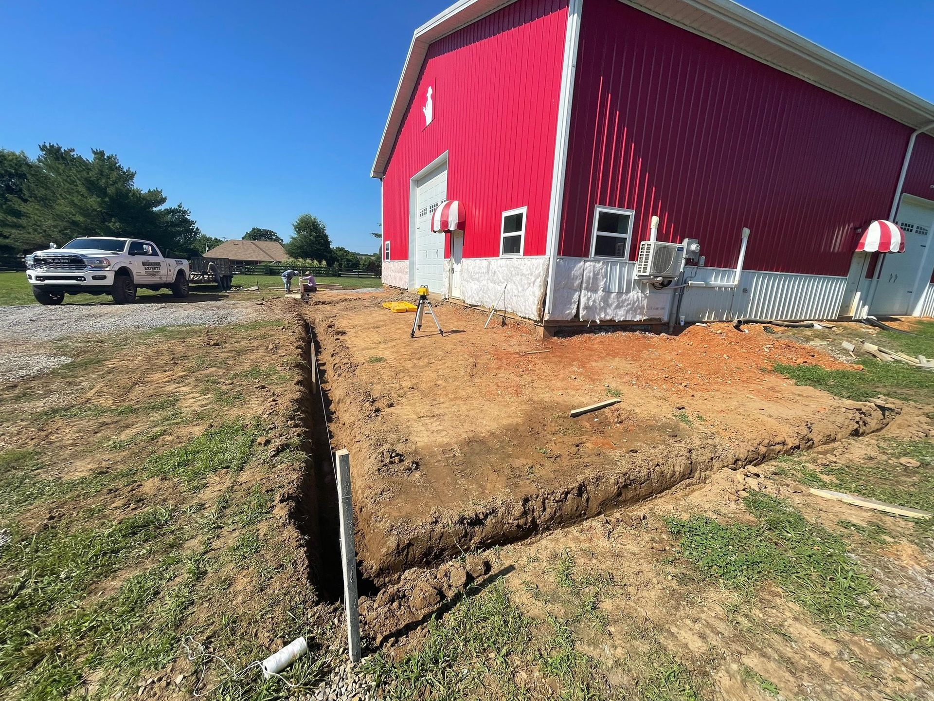 Construction site: Trench dug near a red building with white trim; a truck is in the background.