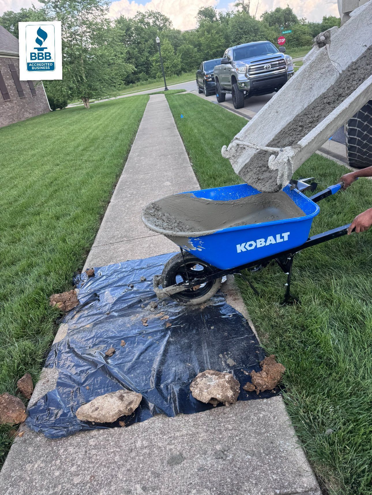 Cement being poured from a truck into a blue wheelbarrow on a sidewalk, next to grass and a house.