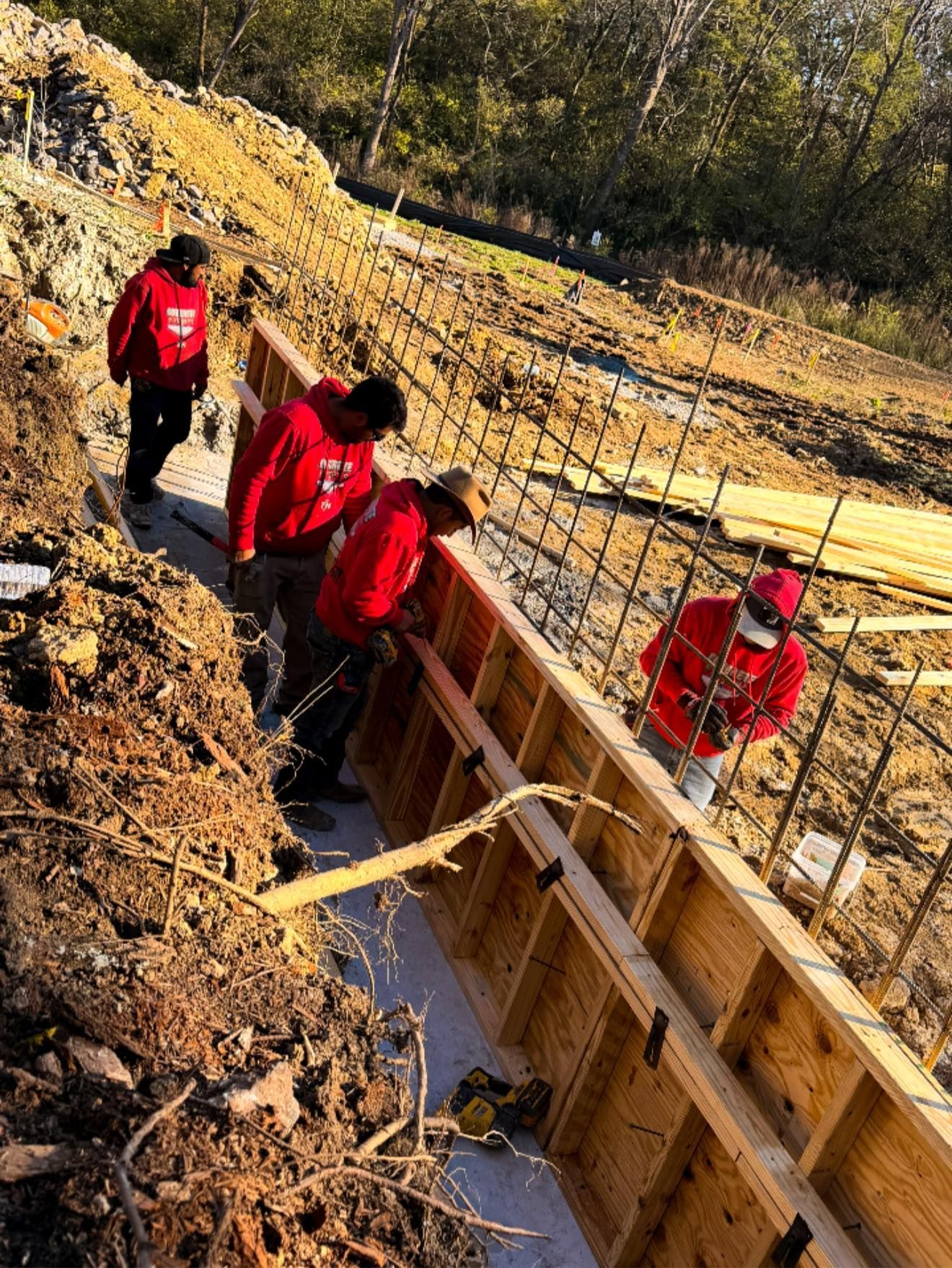 Construction workers in red hoodies build a concrete form, near a dirt embankment.