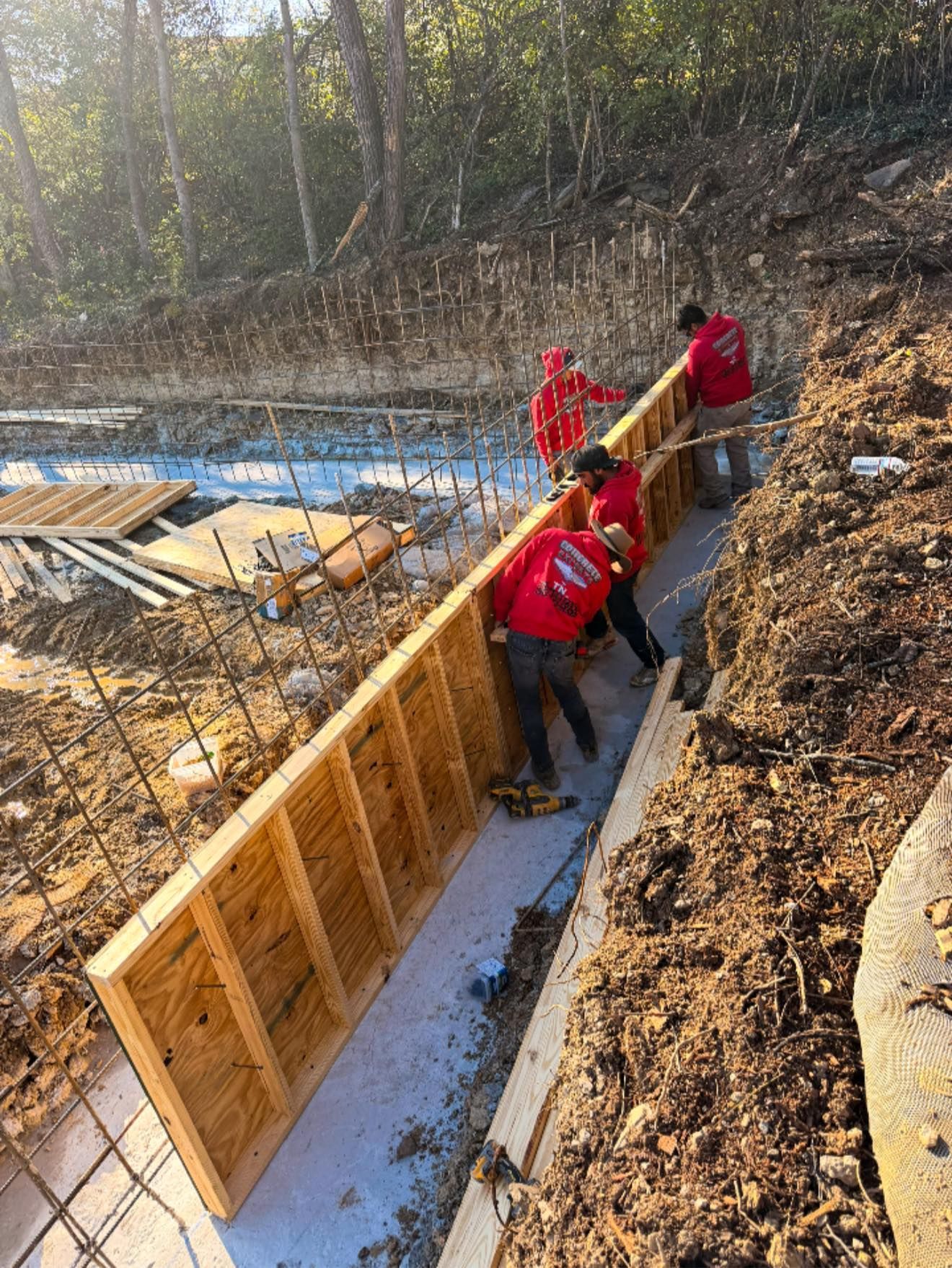Construction workers in red shirts setting up wooden formwork. Outdoor construction site with dirt and concrete.