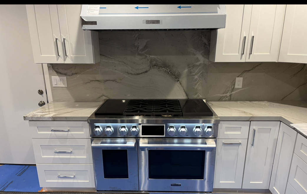 A white kitchen featuring a large stainless steel double oven, gas range, vent hood, and a gray stone-patterned backsplash.