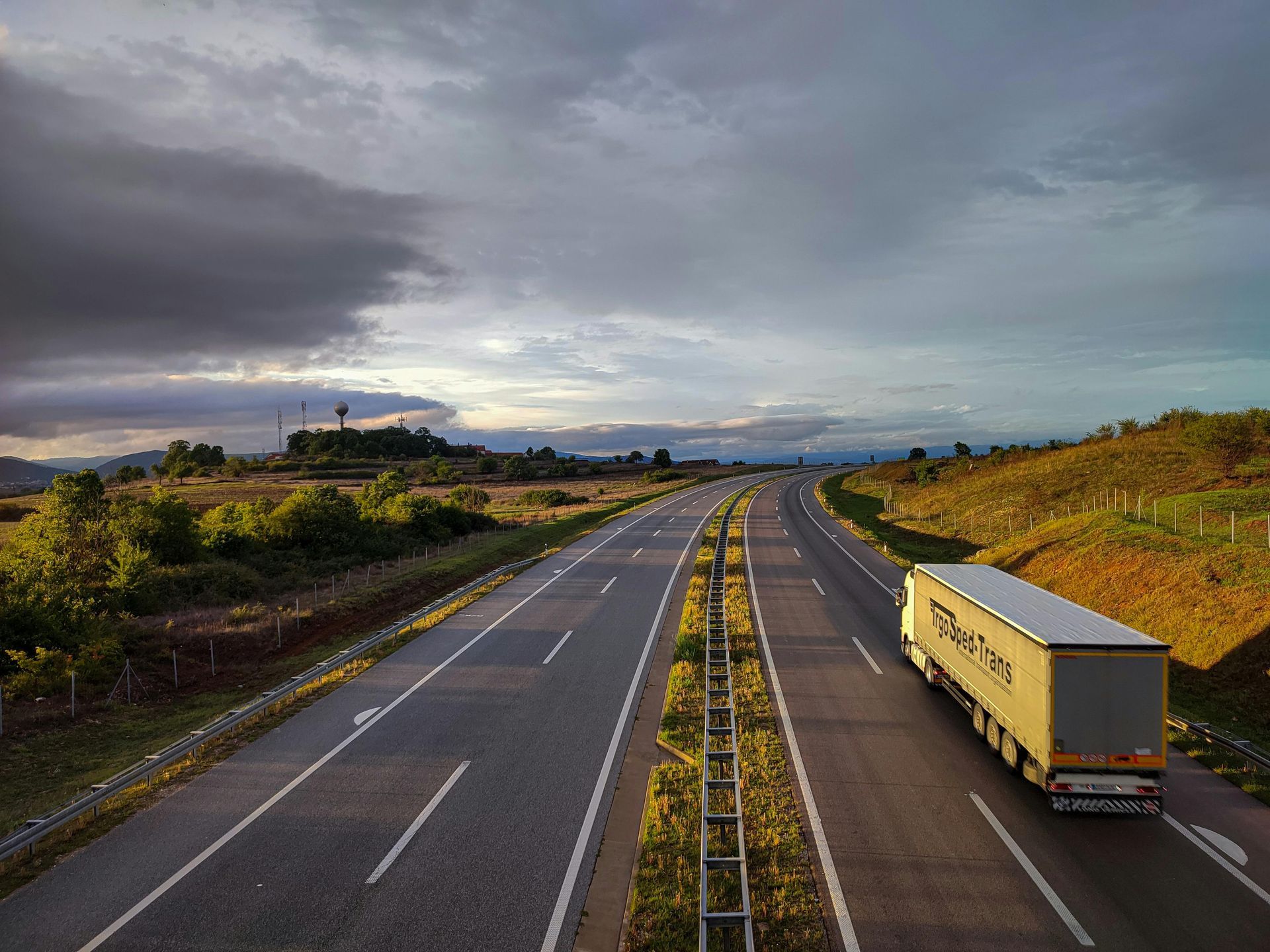 A semi-truck travels on a multi-lane highway through a rural, hilly landscape under a cloudy sky at sunset.