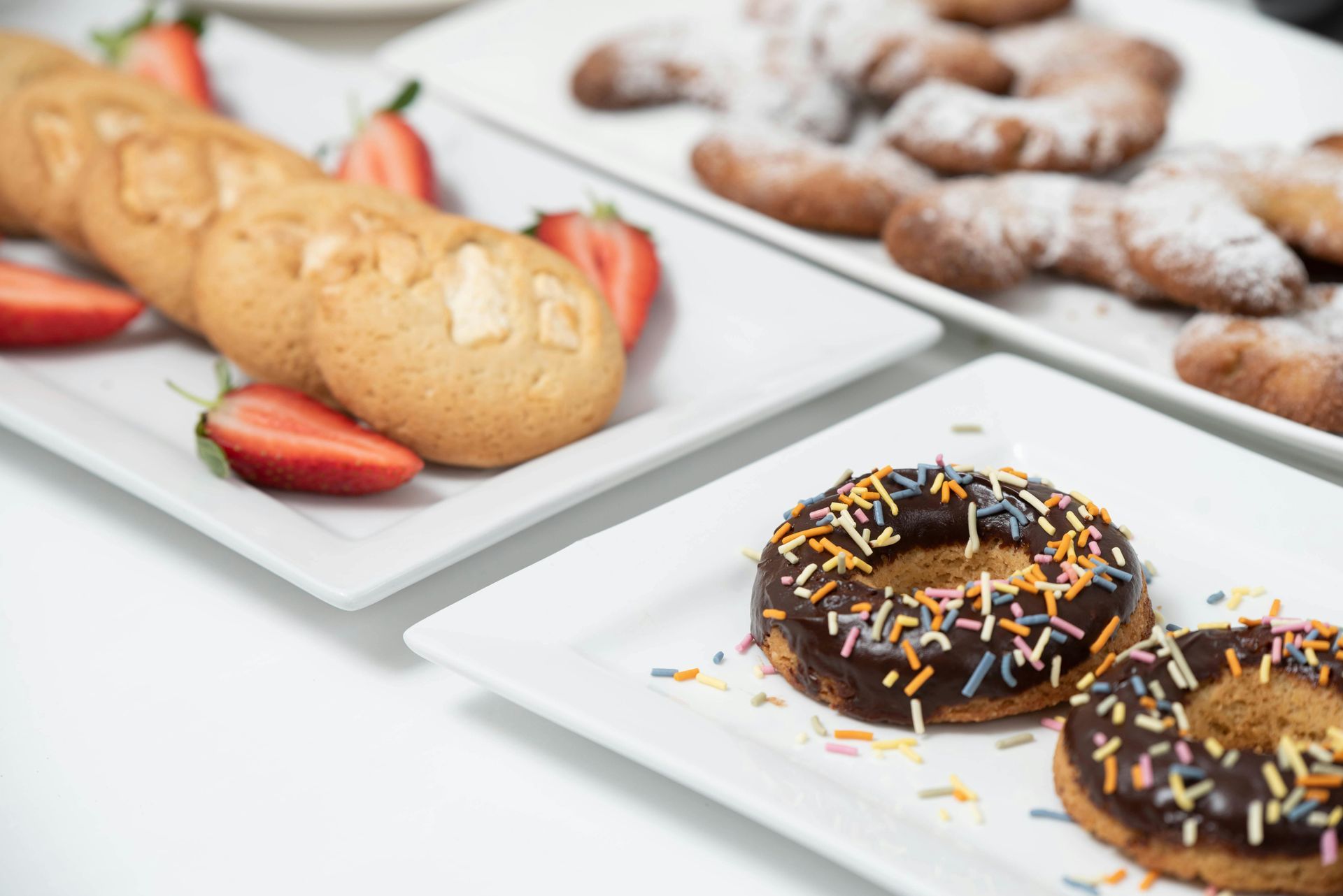 Assorted donuts and cookies on white plates, including chocolate-frosted donuts and pastry rings with strawberries.
