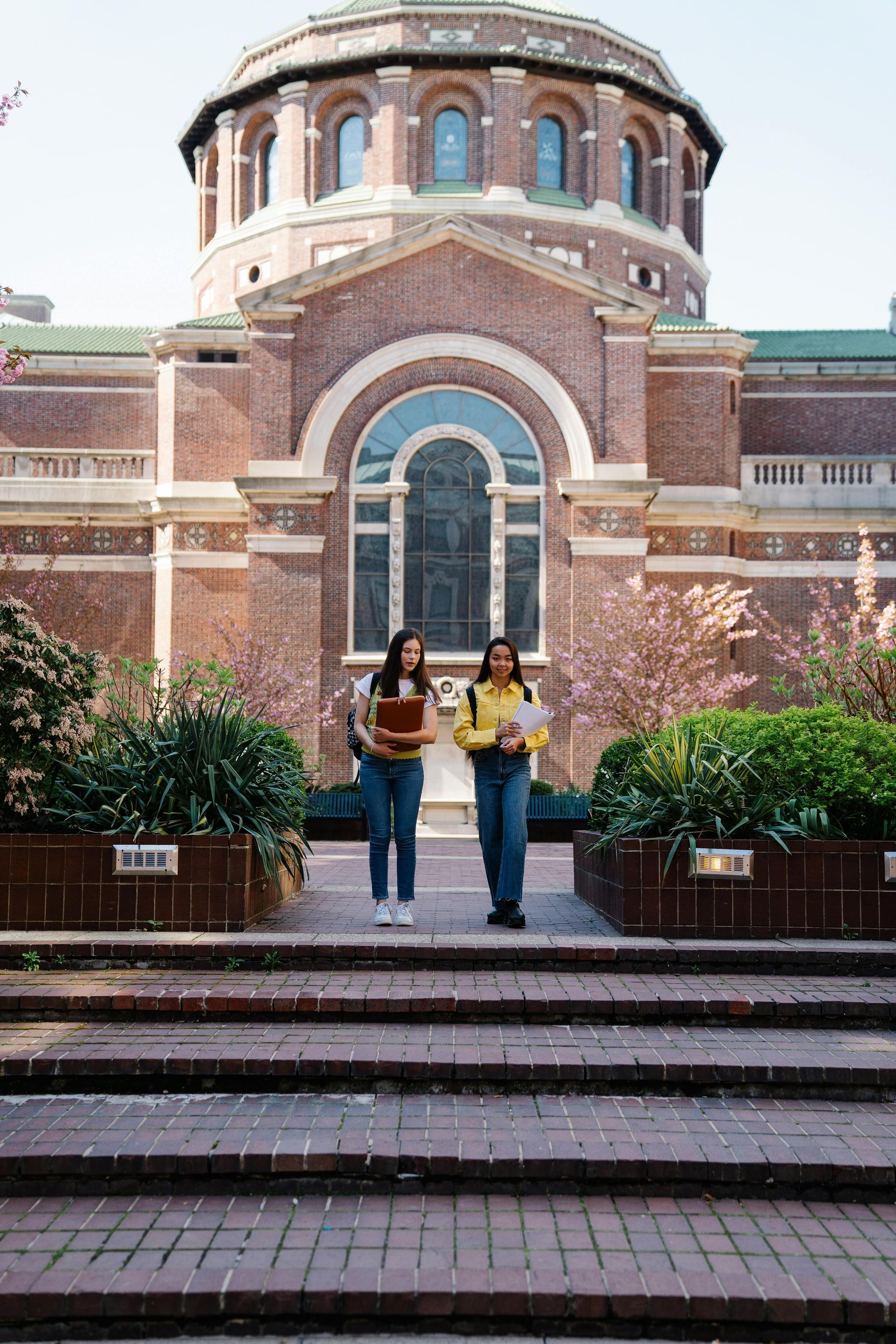 Two students walk up brick steps toward a large, historic brick building with a dome and arched windows.