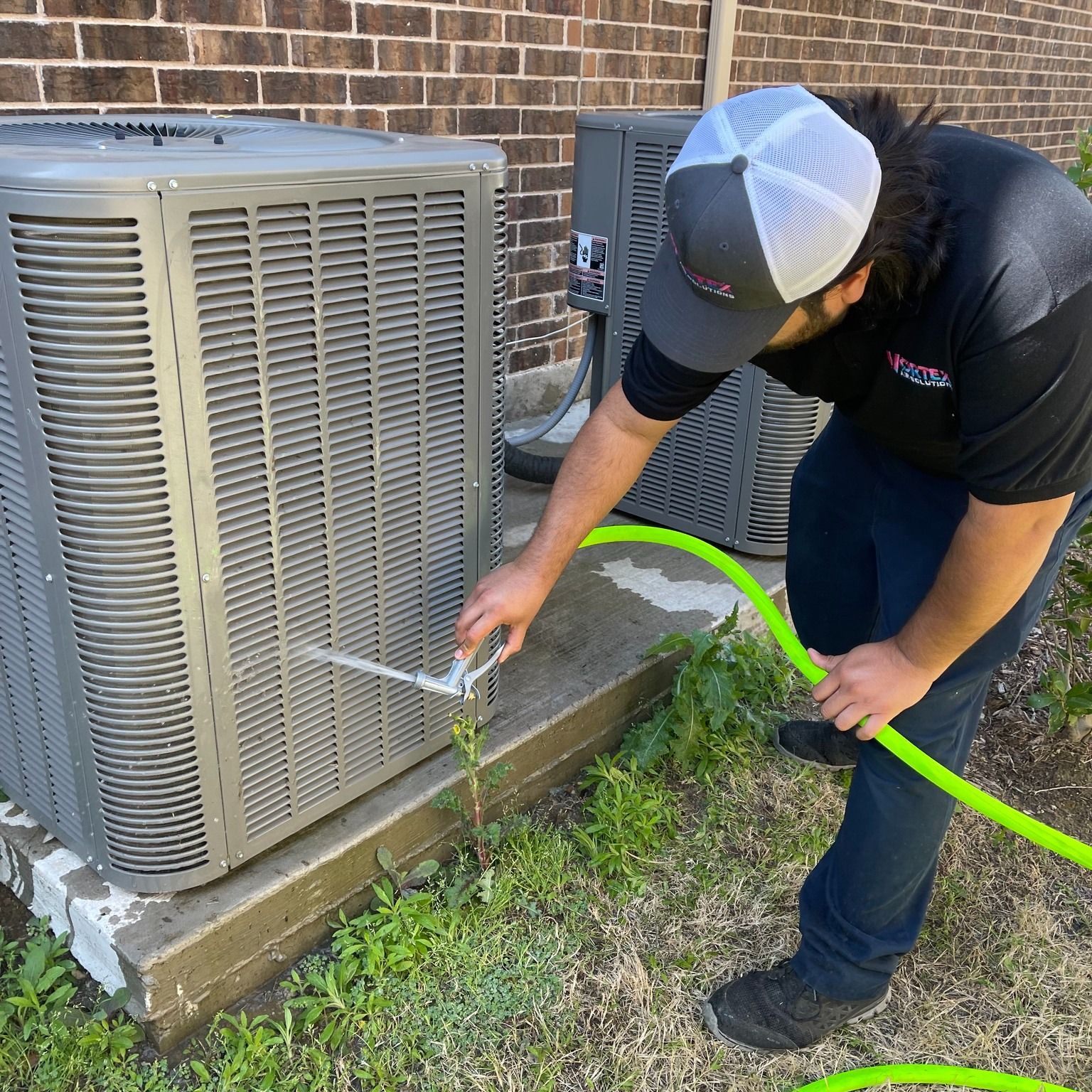 Man cleaning an AC unit with a hose. Gray unit, green hose, wearing a hat, outside near a brick wall.