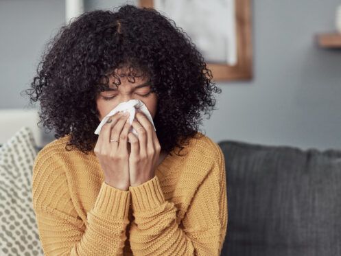 Woman indoors blowing her nose with a tissue; she's wearing a yellow sweater.