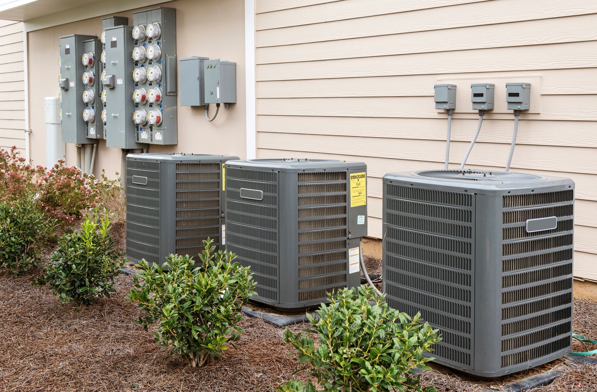 Three air conditioning units outside a building with electrical meters and bushes.
