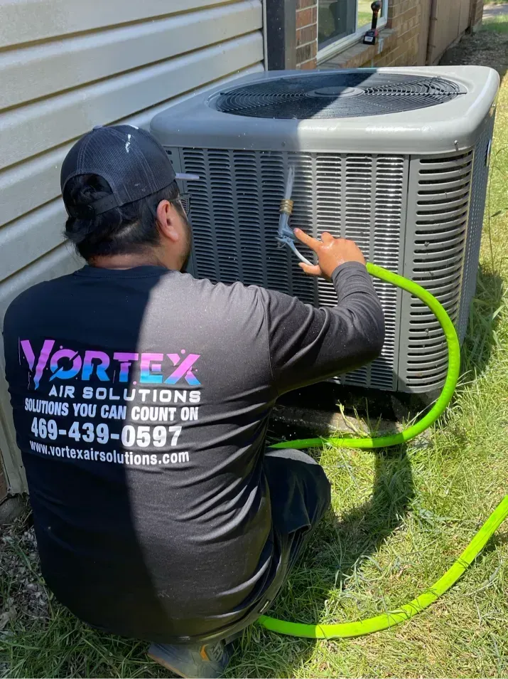 Person in black shirt sprays an AC unit with a green hose outdoors. Logo on shirt: Vortex Air Solutions.