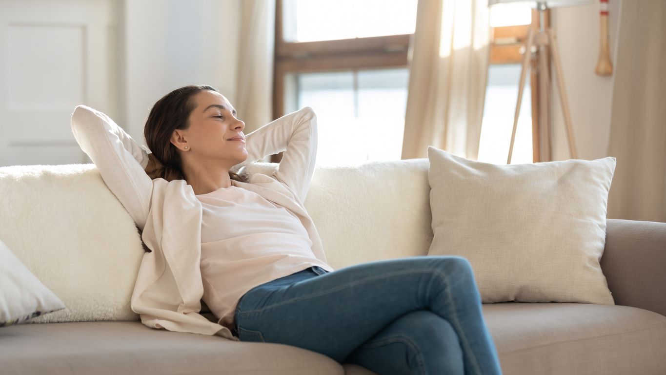 Woman with eyes closed, arms behind her head, relaxing on a couch in a sunlit room.