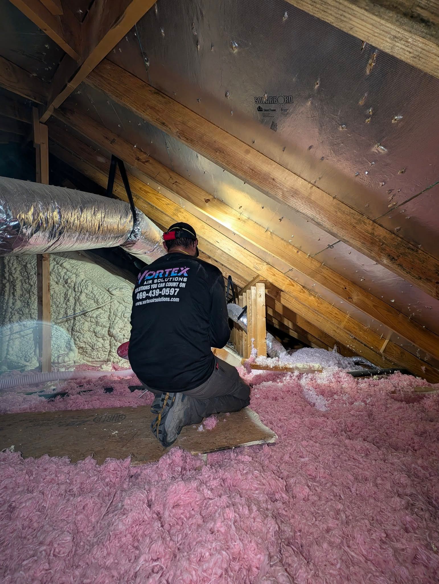 Man in attic inspecting insulation. Pink insulation covers the floor, ductwork visible.
