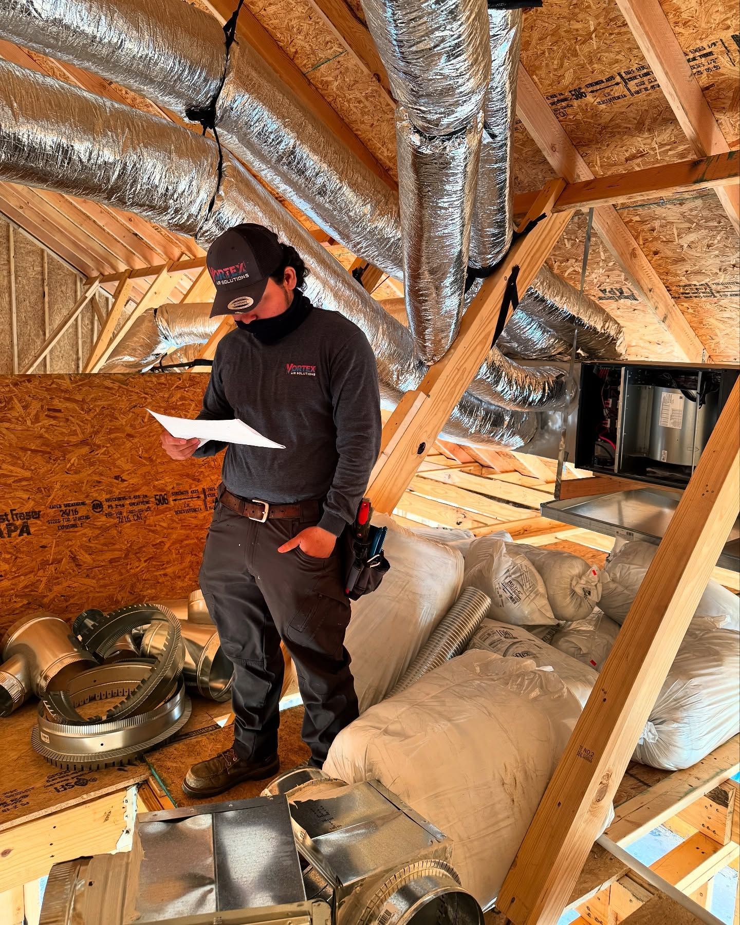 HVAC technician in attic inspecting paperwork, surrounded by ductwork and insulation.