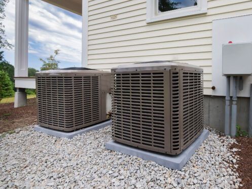 Two outdoor air conditioning units on gravel next to a house with beige siding.