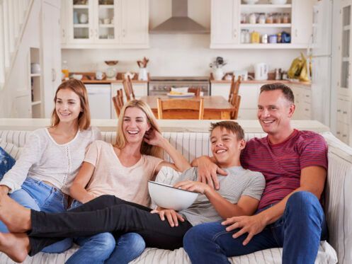 Family watching TV and laughing on a sofa in a living room; bowl of popcorn visible.